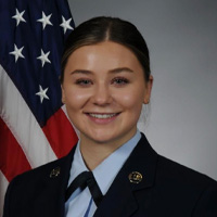 A young woman in a formal military uniform smiles in front of an American flag, posing for an official portrait against a gray background.