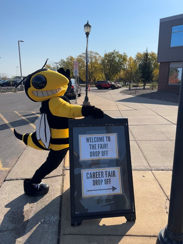 Buzz mascot in yellow and black bee mascot costume stands next to a sidewalk sign that reads “Welcome to the fair! Drop off” and “Career fair drop off.”
