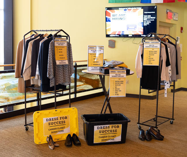 Clothing racks with business attire, shoes, and donation bins labeled Dress for Success Career Closet Donations are displayed indoors. Informational signs and a screen with a We Are Hiring! message are in the background.