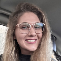 A young woman with long blonde hair and large, light-colored glasses smiles while sitting inside a car. She is wearing a dark top and appears to be happy.