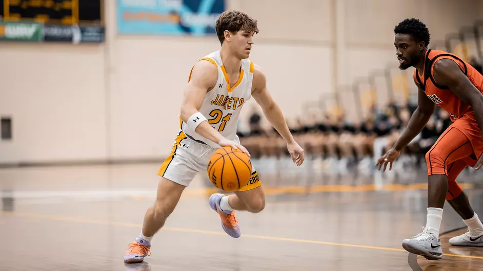 A basketball player in a white and yellow University of Wisconsin-Superior uniform dribbles the ball while being guarded by an opponent in orange and black on an indoor court.