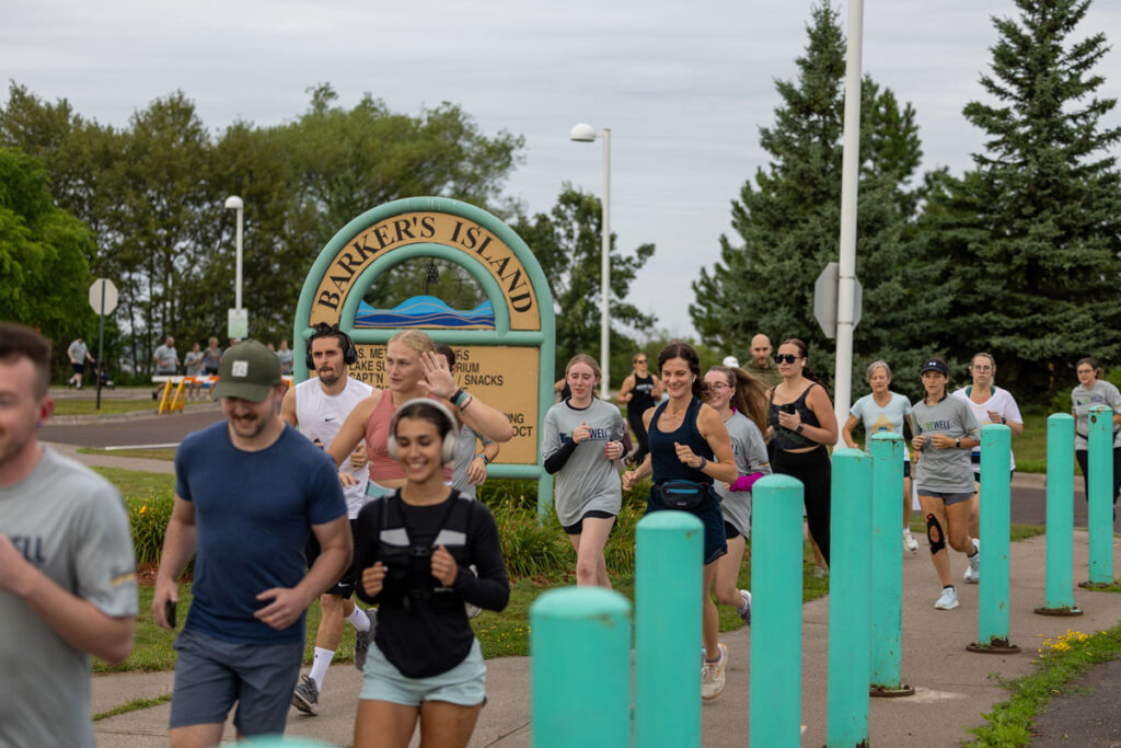 A group of people jog together on a paved path near a sign that reads Barkers Island, participating in a Pruitt Center for Mindfulness and Well-Being event. The scene is outdoors with green trees and overcast skies in the background.