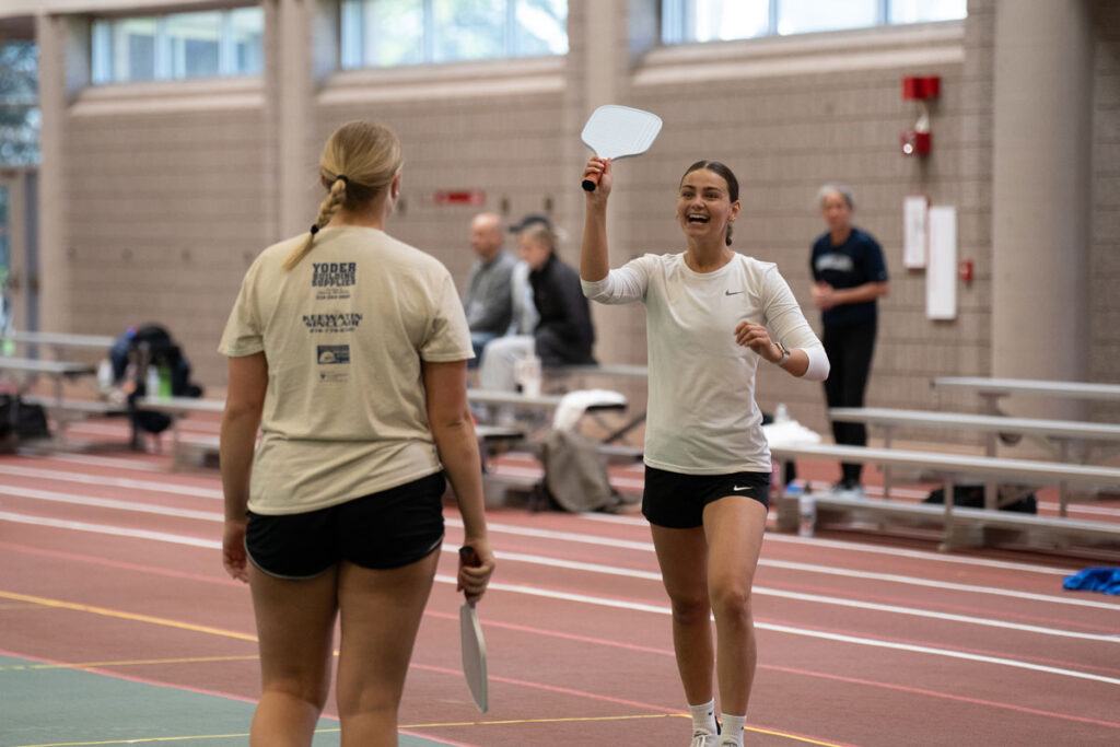 Two women play pickleball indoors at the Pruitt Center for Mindfulness and Well-Being; one looks excited and raises her paddle while smiling at her opponent. The court has a green and red surface, with benches and spectators in the background.