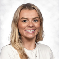 A woman with long blonde hair and light skin smiles at the camera. She is wearing a white top and a necklace, posed against a light, softly blurred background.