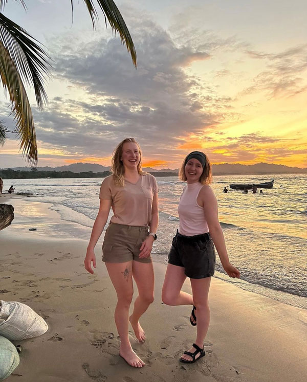 Two women smiling and posing on a sandy beach at sunset, with palm leaves overhead, gentle waves, and a boat in the background—capturing the adventure of a Study Away at UW-Superior.