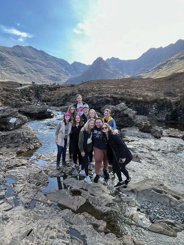 A group of ten people, part of Study Away at UW-Superior, poses and smiles on rocky terrain by a stream, with mountains and a bright sky in the background. Some wear jackets and sunglasses, suggesting cool weather.